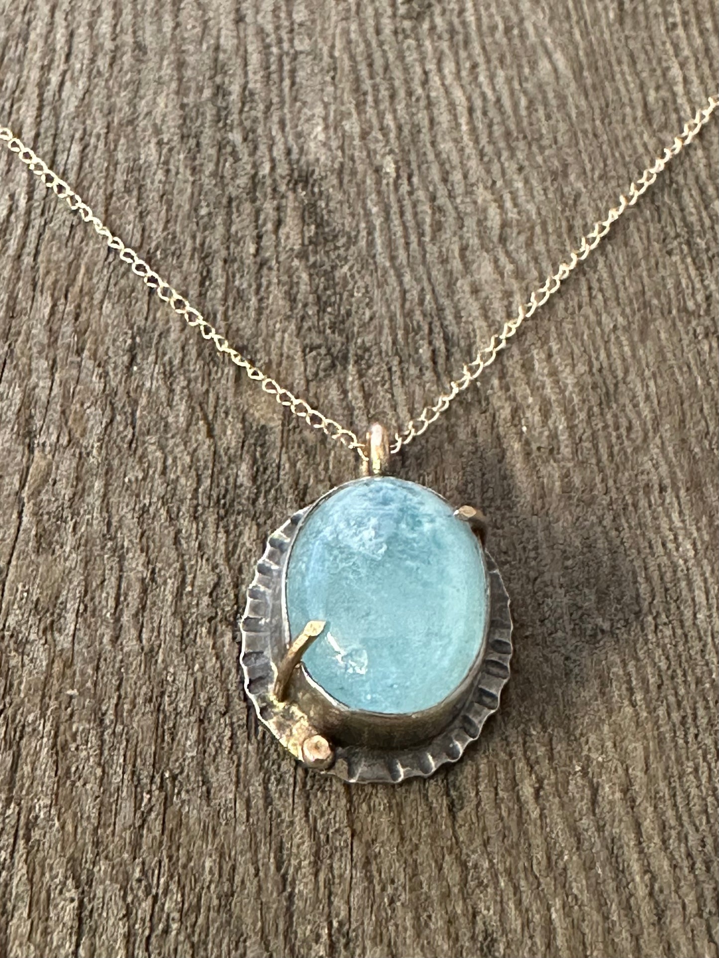 Blue Aquamarine oval stone set in silver with gold prongs and a small gold ball in the lower left corner on a gold chain laying on a wooden background.