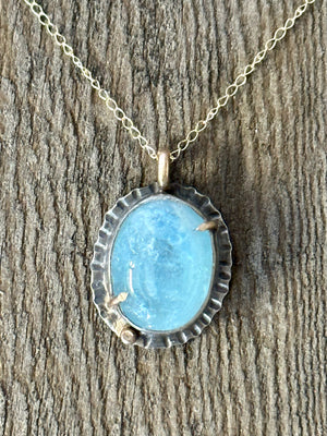 Blue Aquamarine oval stone set in silver with gold prongs and a small gold ball in the lower left corner on a gold chain laying on a wooden background.