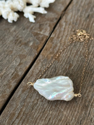 Large white cloud shaped pearl on a gold chain with coral in the upper left corner on a wood background