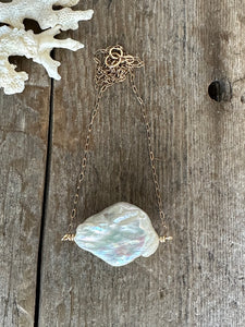 Large white cloud shaped pearl on a gold chain with coral in the upper left corner on a wood background