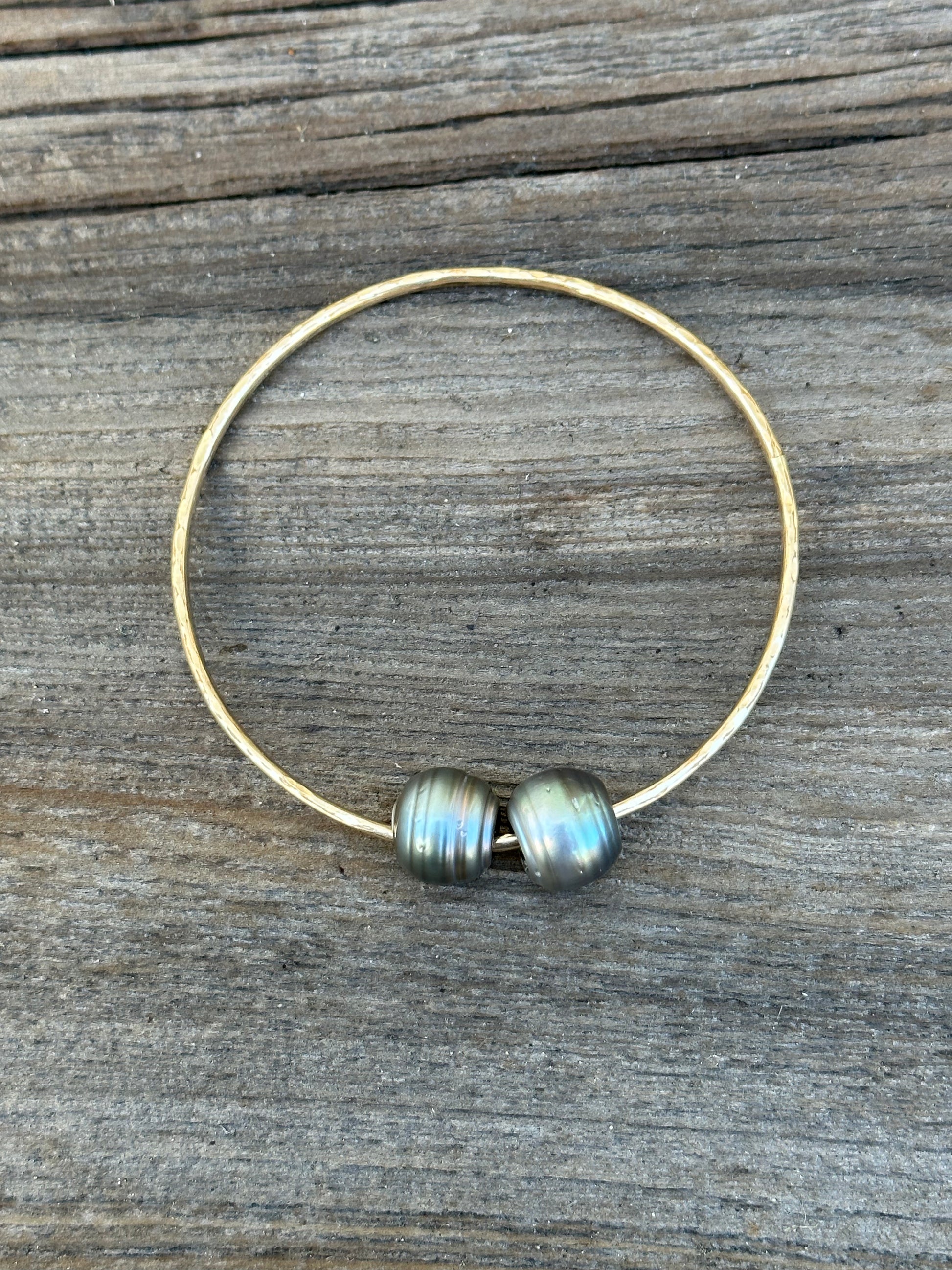 Two black baroque Tahitian pearls on a gold bangle resting on a wooden background