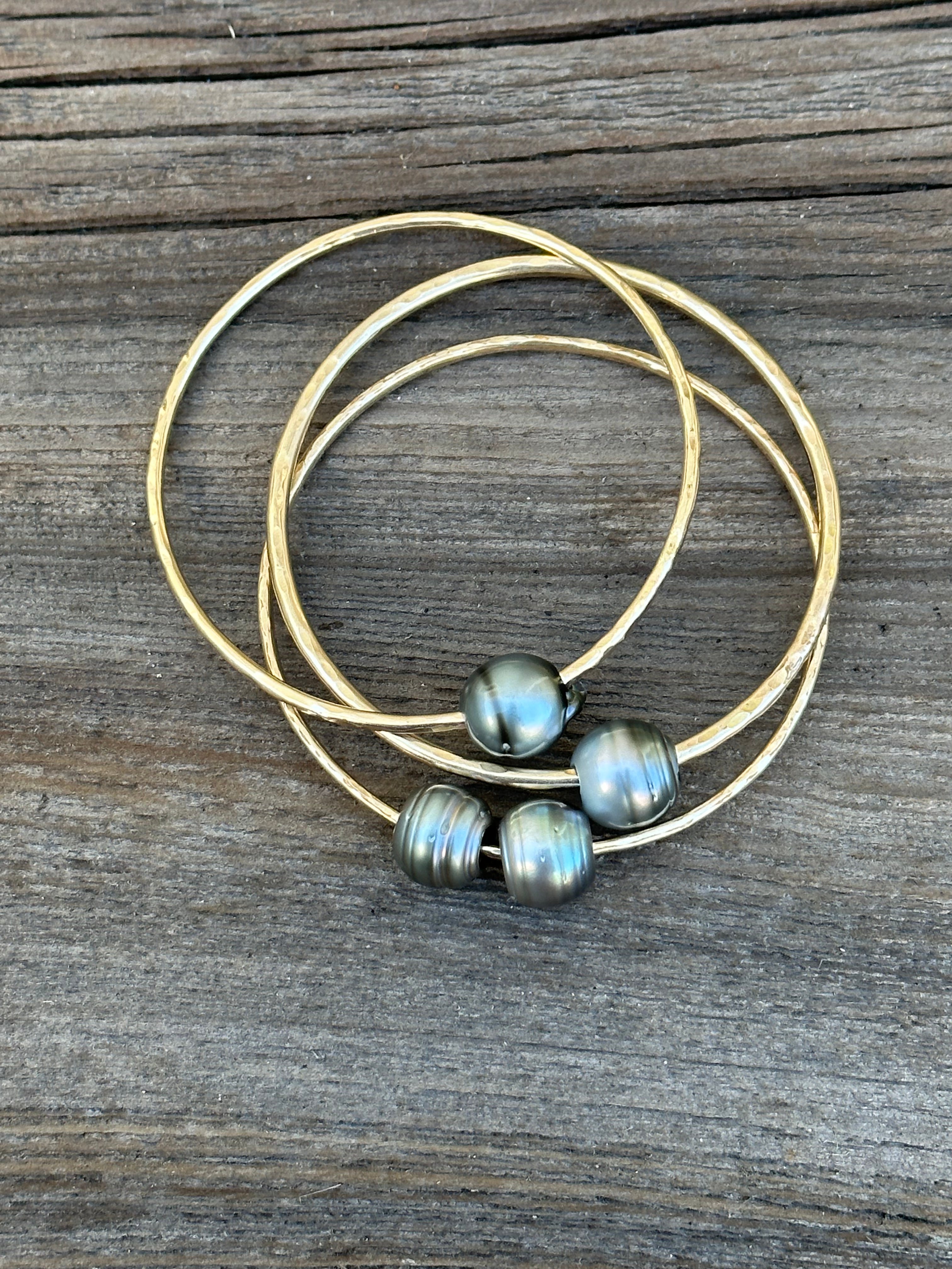 Three gold bangles with Tahitian pearls stacked on top of each other on a wood background.