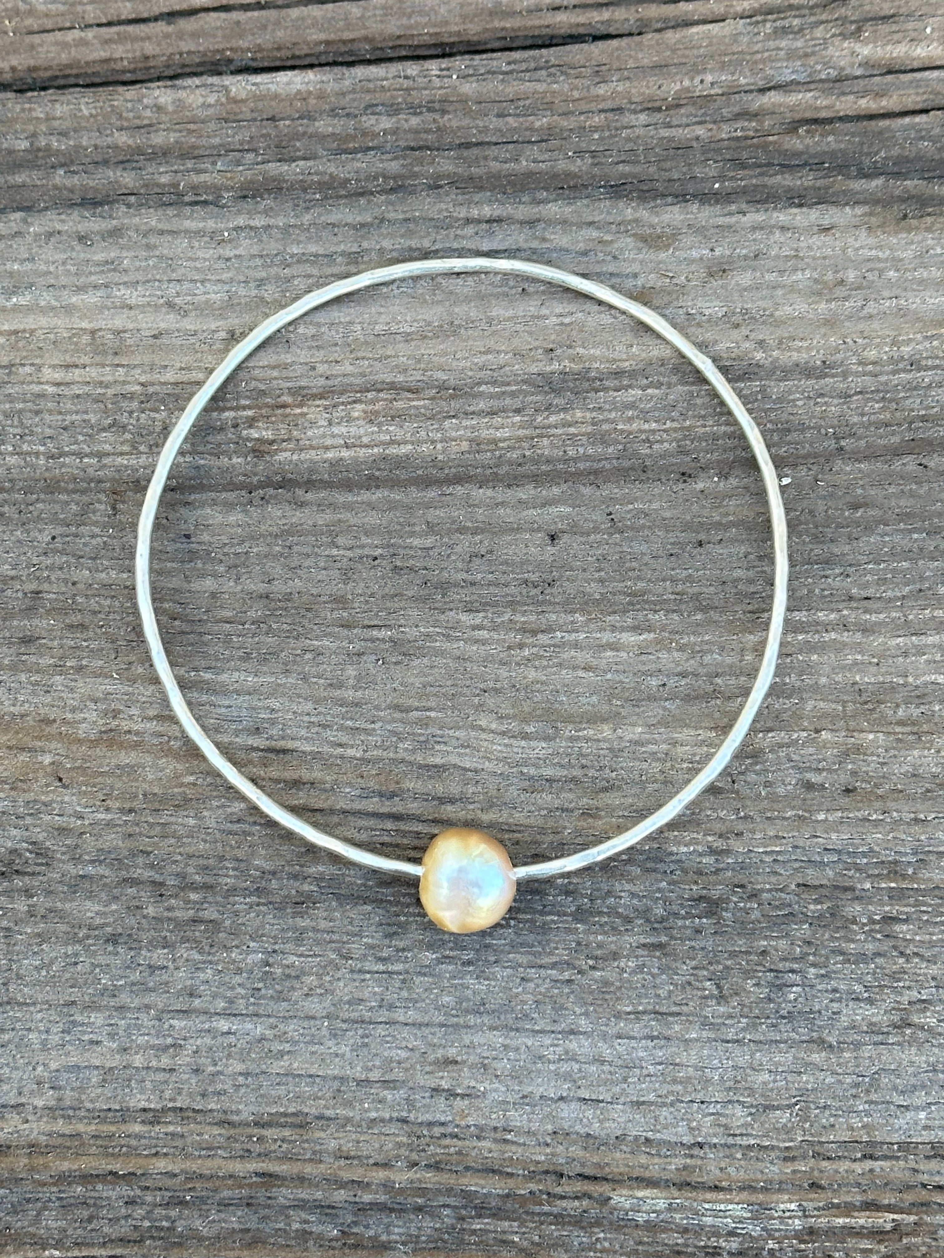 A single silver hammered bangle with a golden pearl on a wooden background.