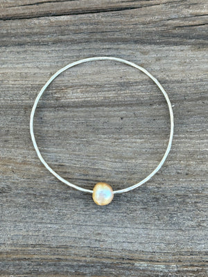 A single silver hammered bangle with a golden pearl on a wooden background.