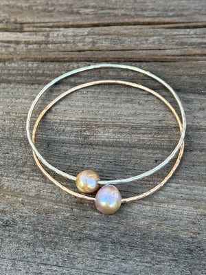 Two bangles with hammered wire, one silver, one gold laying on a wooden background. Both have a small golden freshwater pearl on them.