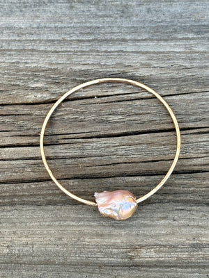 A gold bangle with a large oddly shaped fireball pearl in light hues of pink on a wooden background.