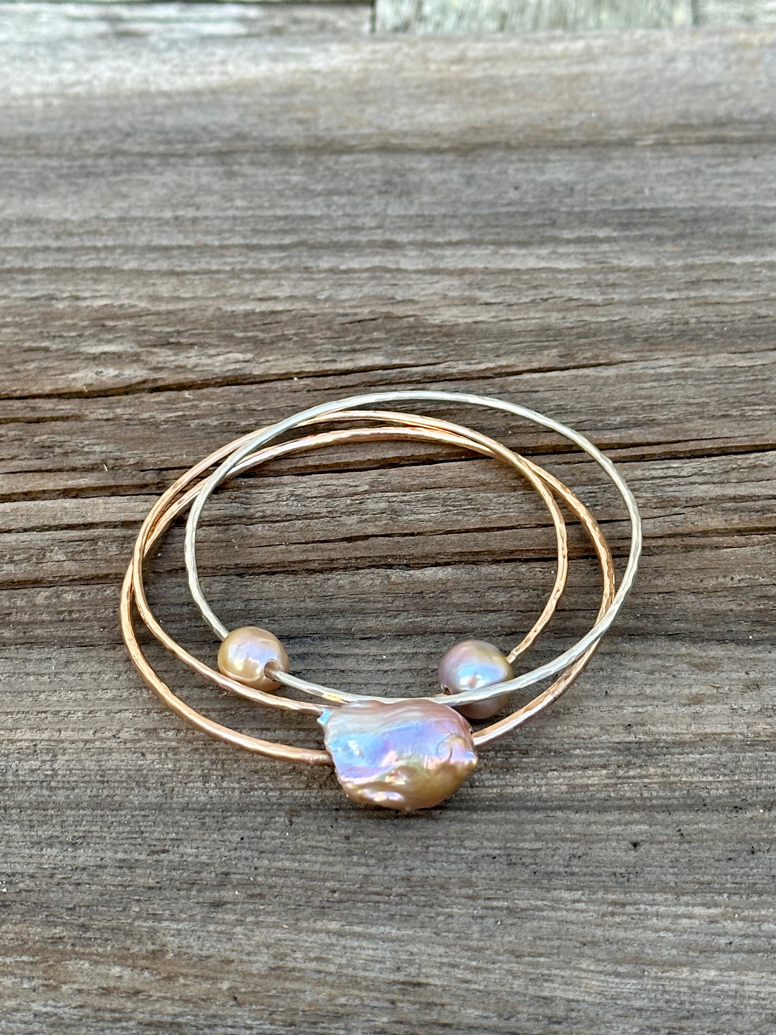 Three gold bangles one with a large oddly shaped fireball pearl in light pink and two with smaller pink pearls on a wooden background.