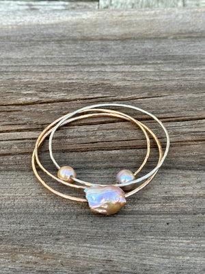 Three gold bangles one with a large oddly shaped fireball pearl in light pink and two with smaller pink pearls on a wooden background.