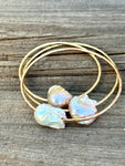 Three gold bangles with large oddly shaped fireball pearls in light hues of pink and peach and white on a wooden background.
