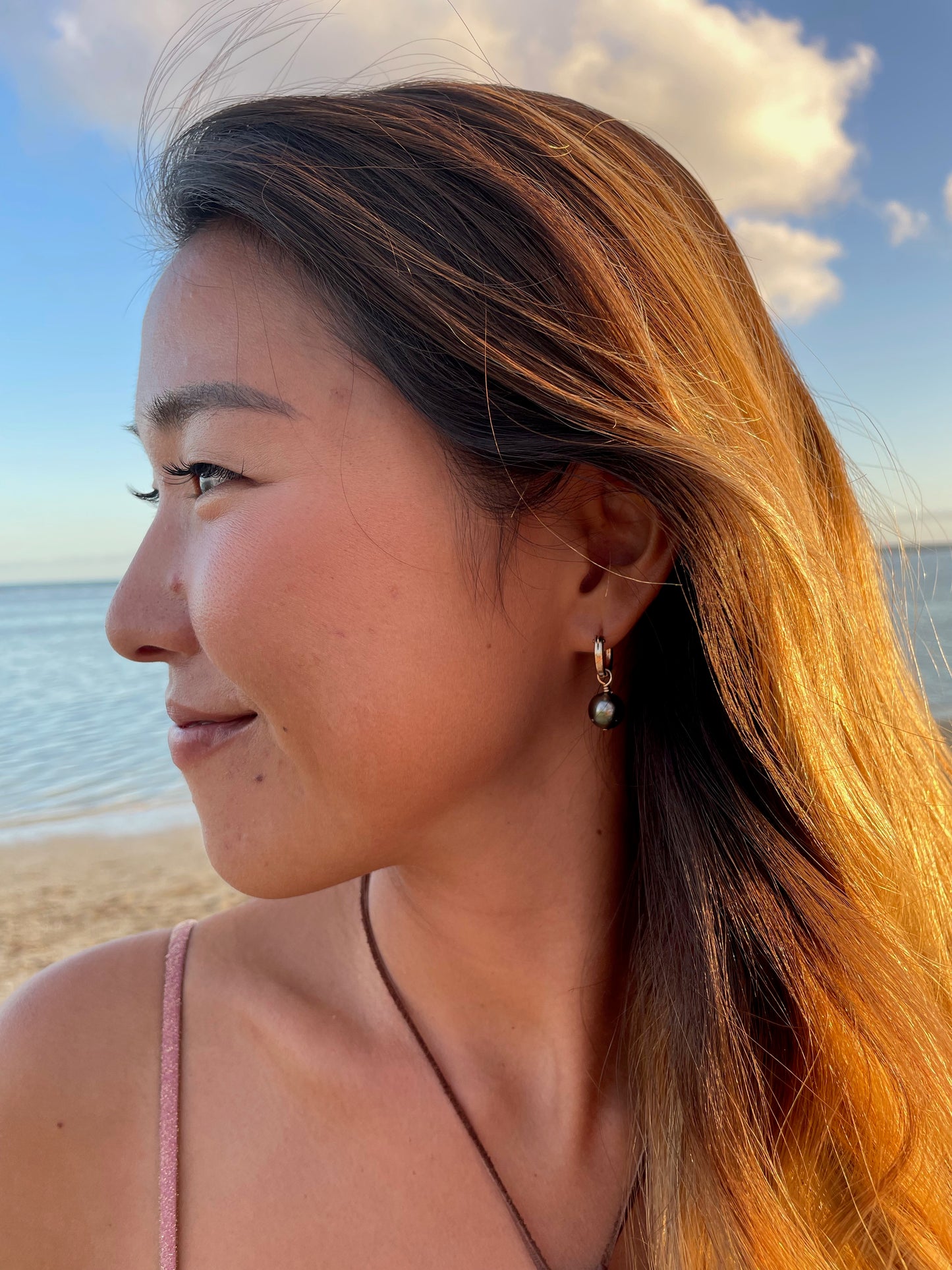 a pretty girl infront of the ocean looking off in the distance. she is wearing a little gold hoop with a black pearl dangliing on it