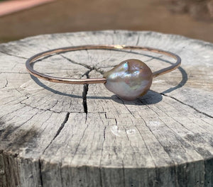 A gold bangle with a large oddly shaped fireball pearl in light hues of pink on a wooden stump background.