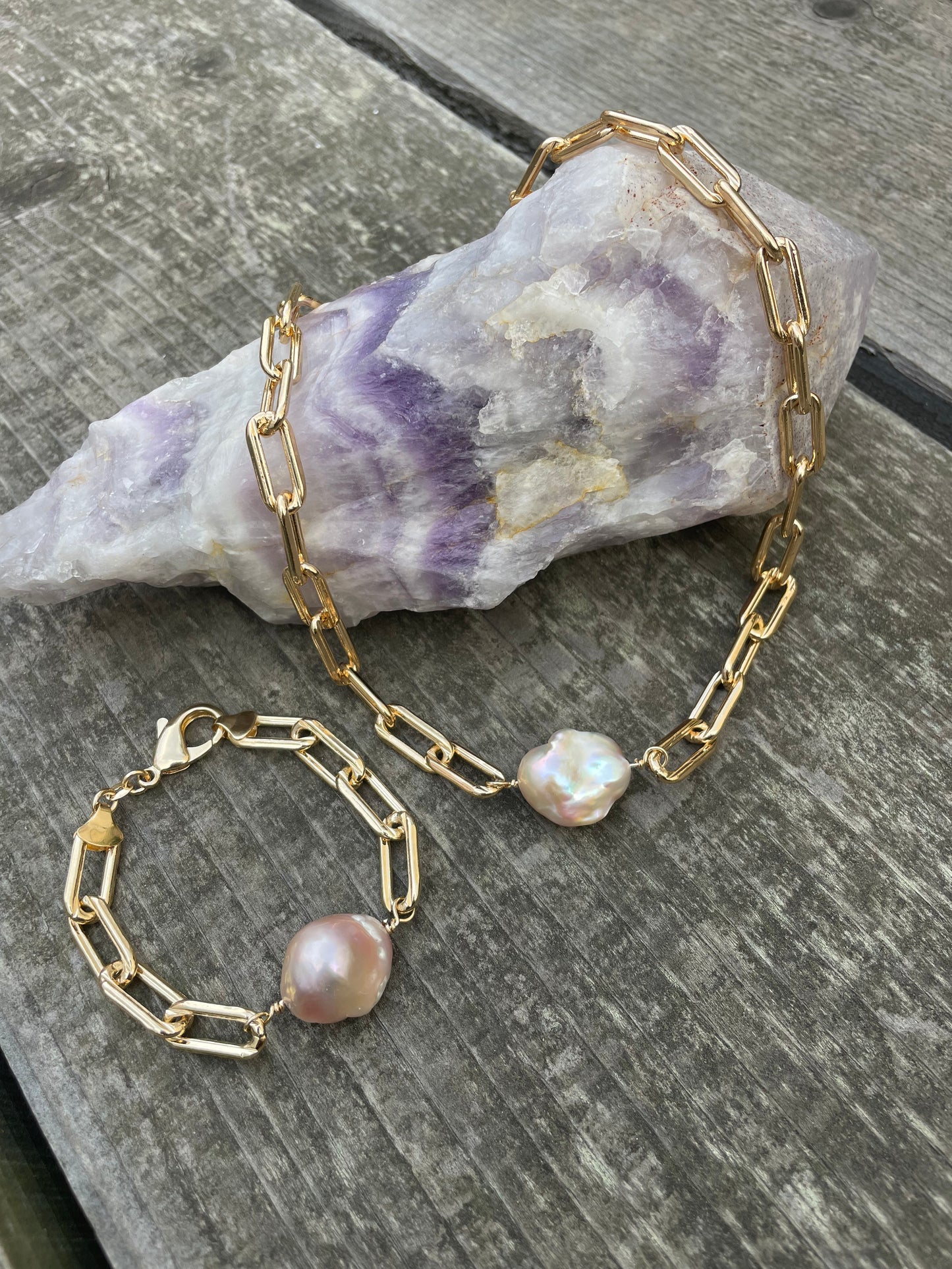 A large purple crystal with a bracelet and necklace draped off of it. Both are large link gold chains with a single large baroque pearl in the center on a wooden background.