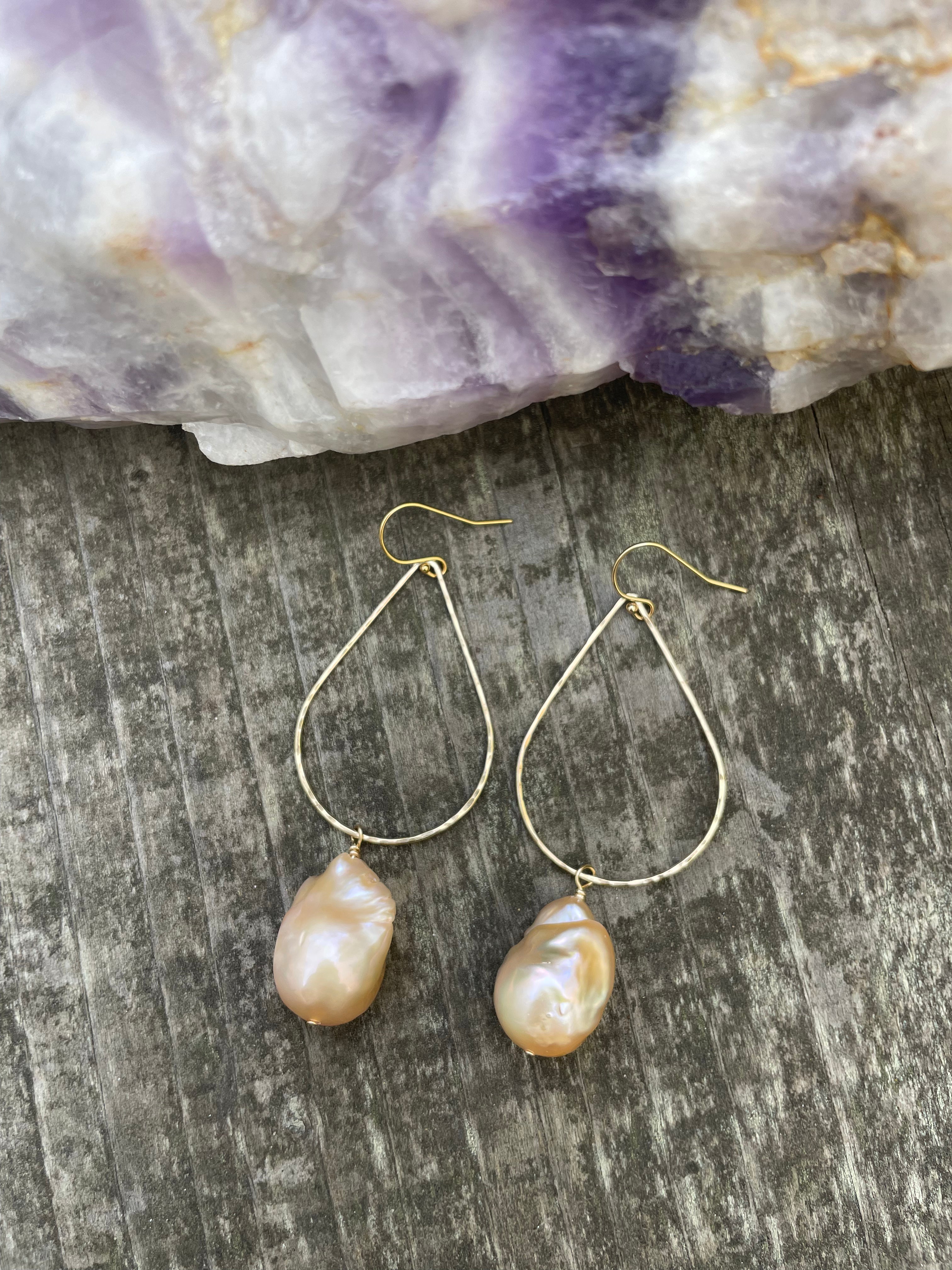 Large pink baroque fireball pearls hanging off of a teardrop shaped gold wire with ear wires on a wooden background. Above the earrings is a purple striped crystal.