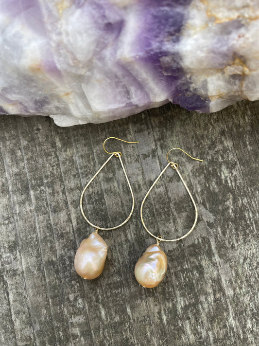 Large pink baroque fireball pearls hanging off of a teardrop shaped gold wire with ear wires on a wooden background. Above the earrings is a purple striped crystal.