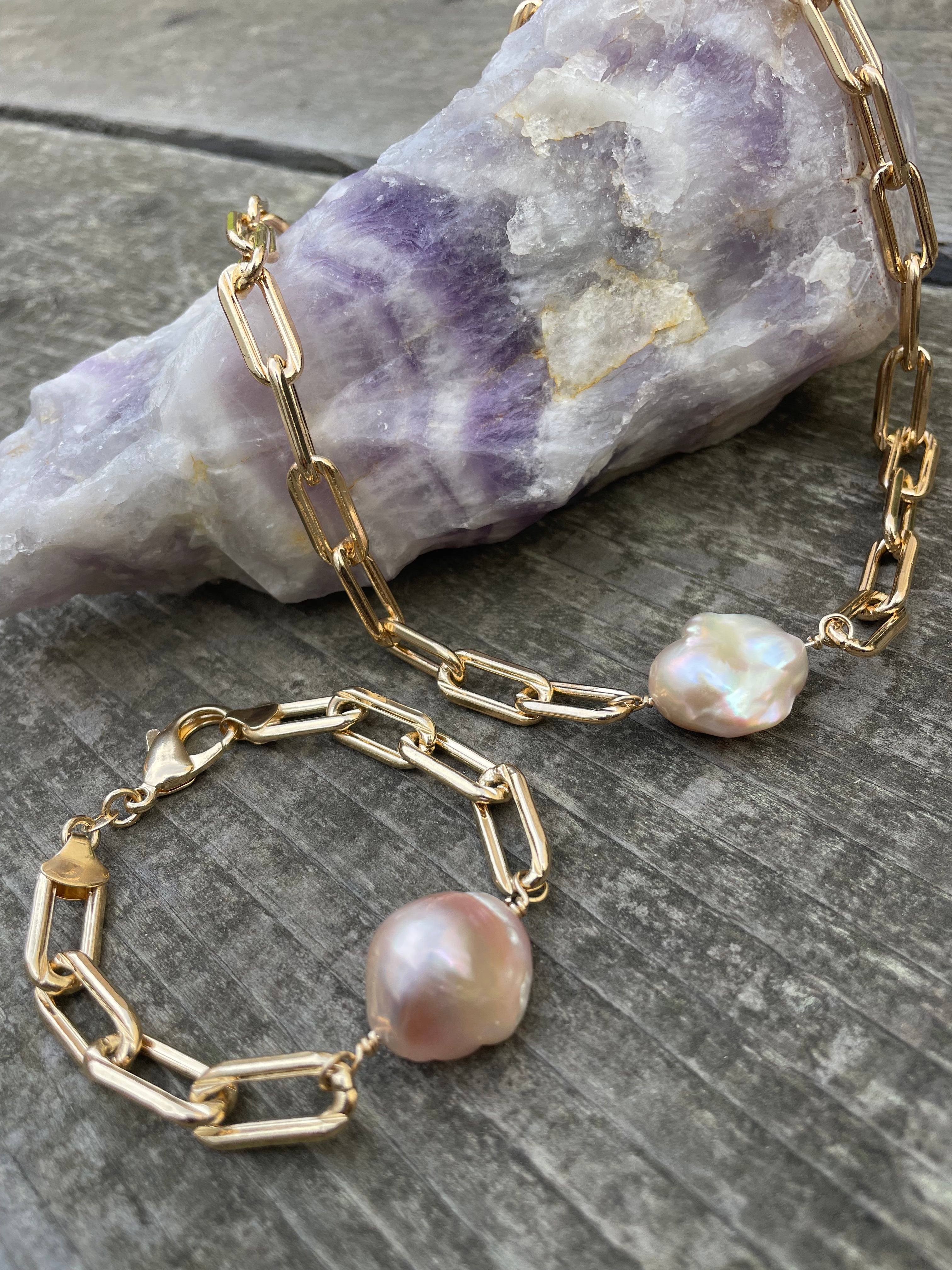 two extra large gold linked chains with large pink fireball shaped pearls laying on a purple crystal on a wooden background. one is a bracelet and the other is a necklace.