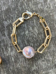 large gold linked bracelet with a large pink baroque pearl on a wooden background.