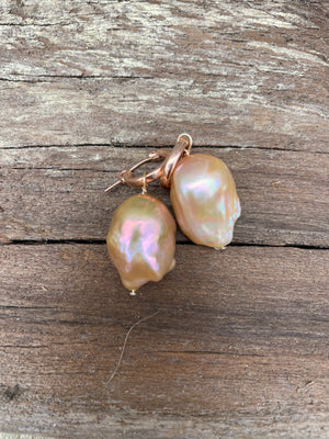 A pair of oddly shaped pinkish pearls on small rose gold lever back hoops laying on a wooden board.
