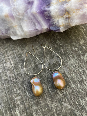 Large bronze baroque fireball pearls hanging off of a teardrop shaped gold wire with ear wires on a wooden background. Above the earrings is a purple striped crystal