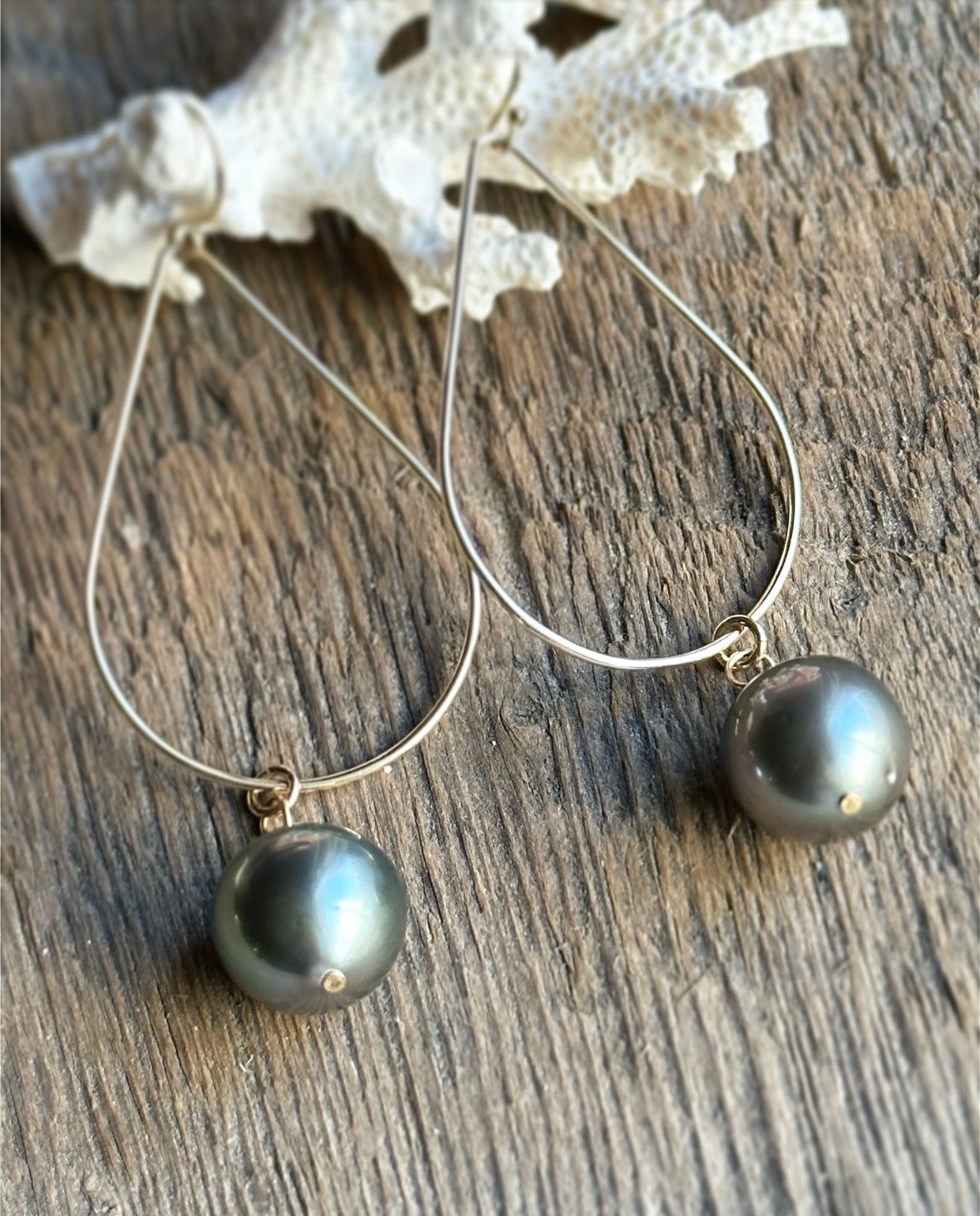 a pair of earrings with black pearls on a golden wire in the shape of ateardrop lays on white coral on a wooden background