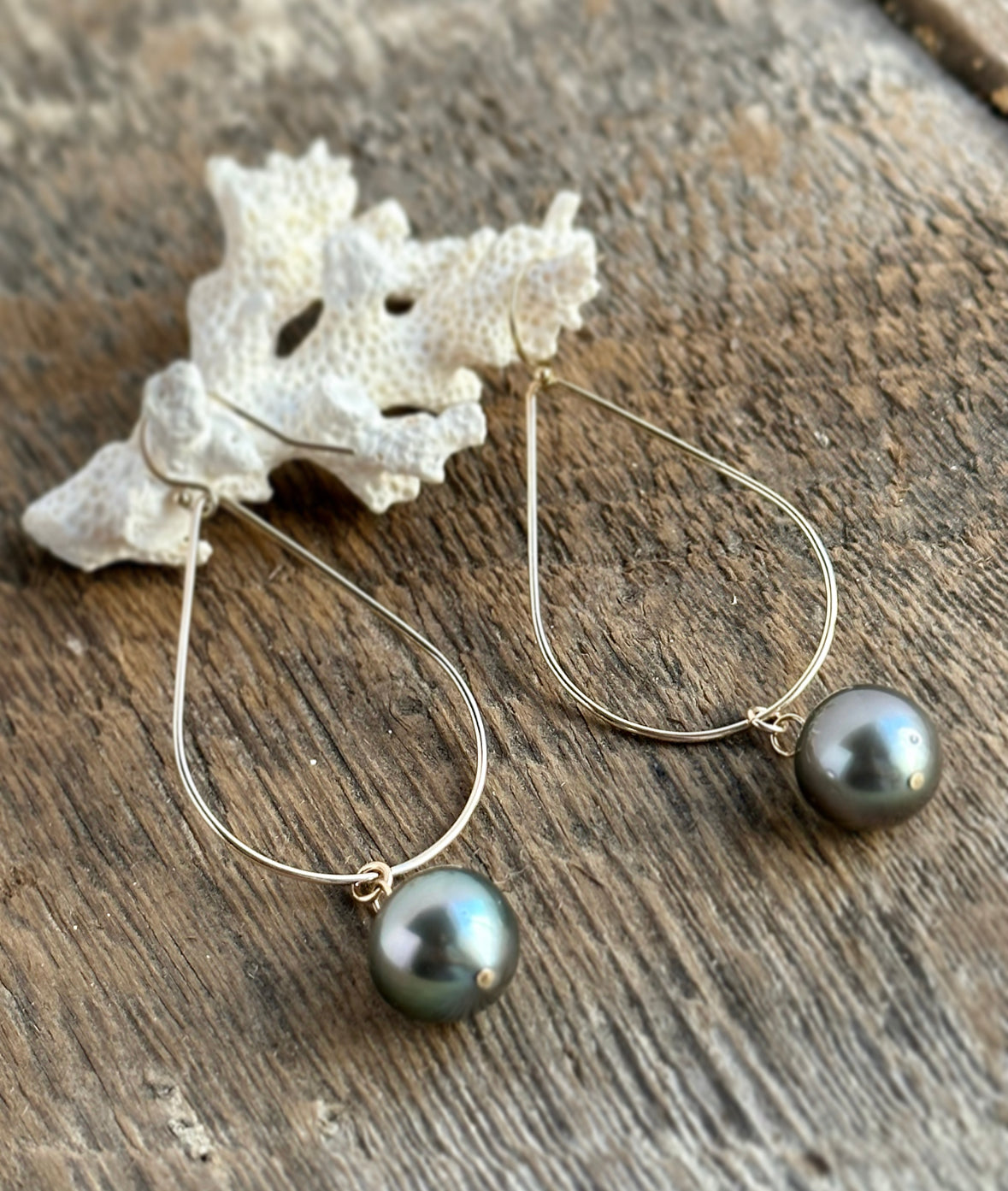 A pair of teardrop hoops in gold with small Tahitian pearls dangling. the earrings are hanging on a piece of coral on a wooden background