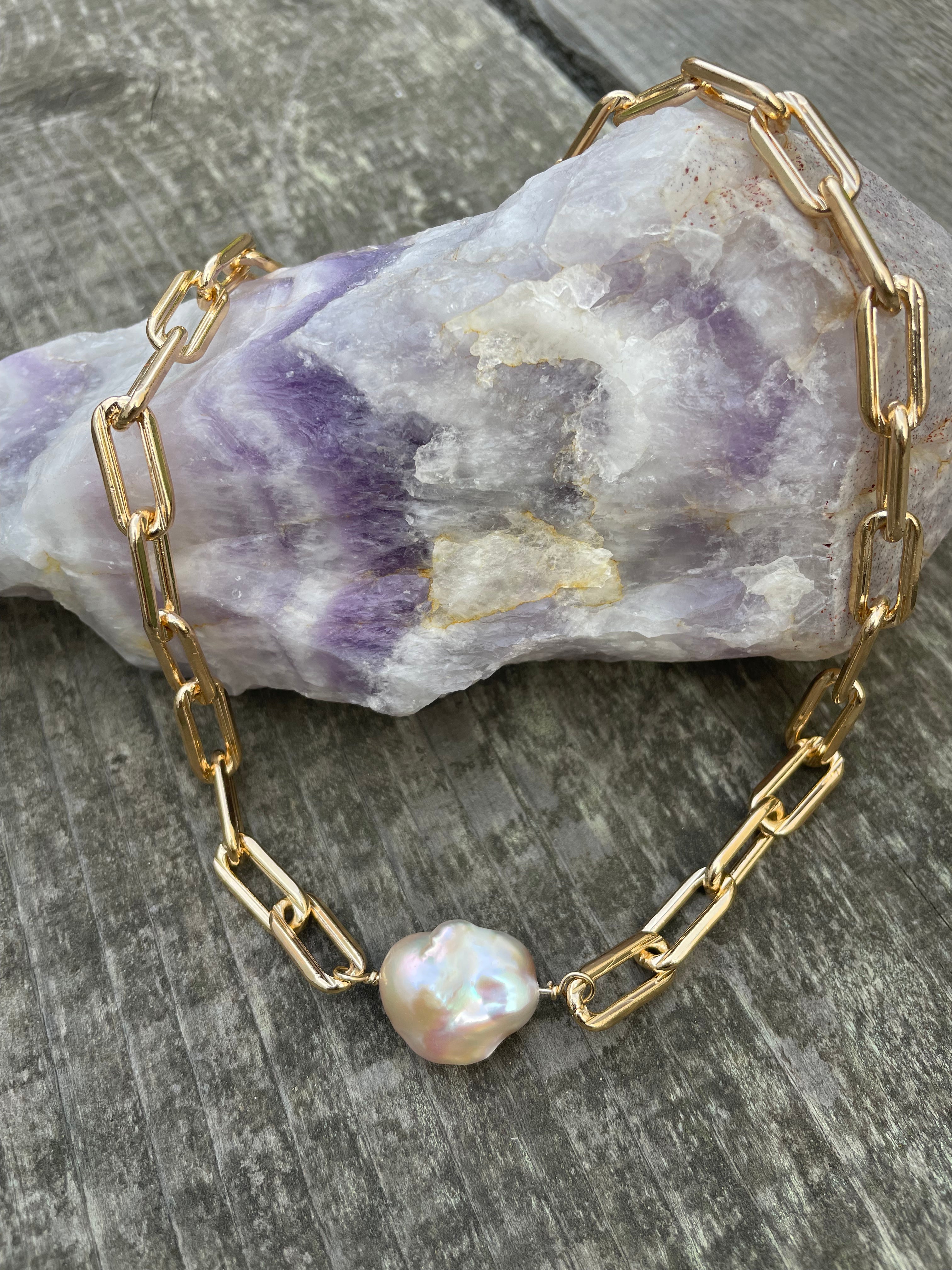 Extra large gold linked chain with a large pink fireball shaped pearl laying on a purple crystal on a wooden background