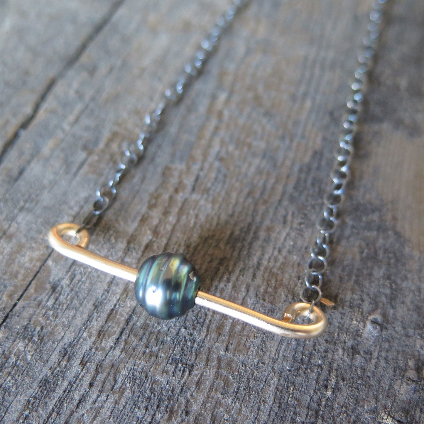 A necklace on a wooden background, there is a gold bar going through a balck pearl and suspended on a sterling silver chain that has been darkened to a grey.