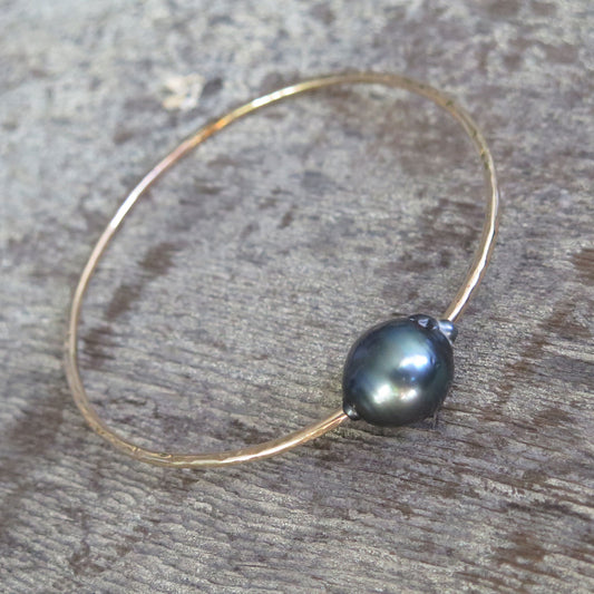 a single black pearl on a yellow gold bangle sits on a wooden background