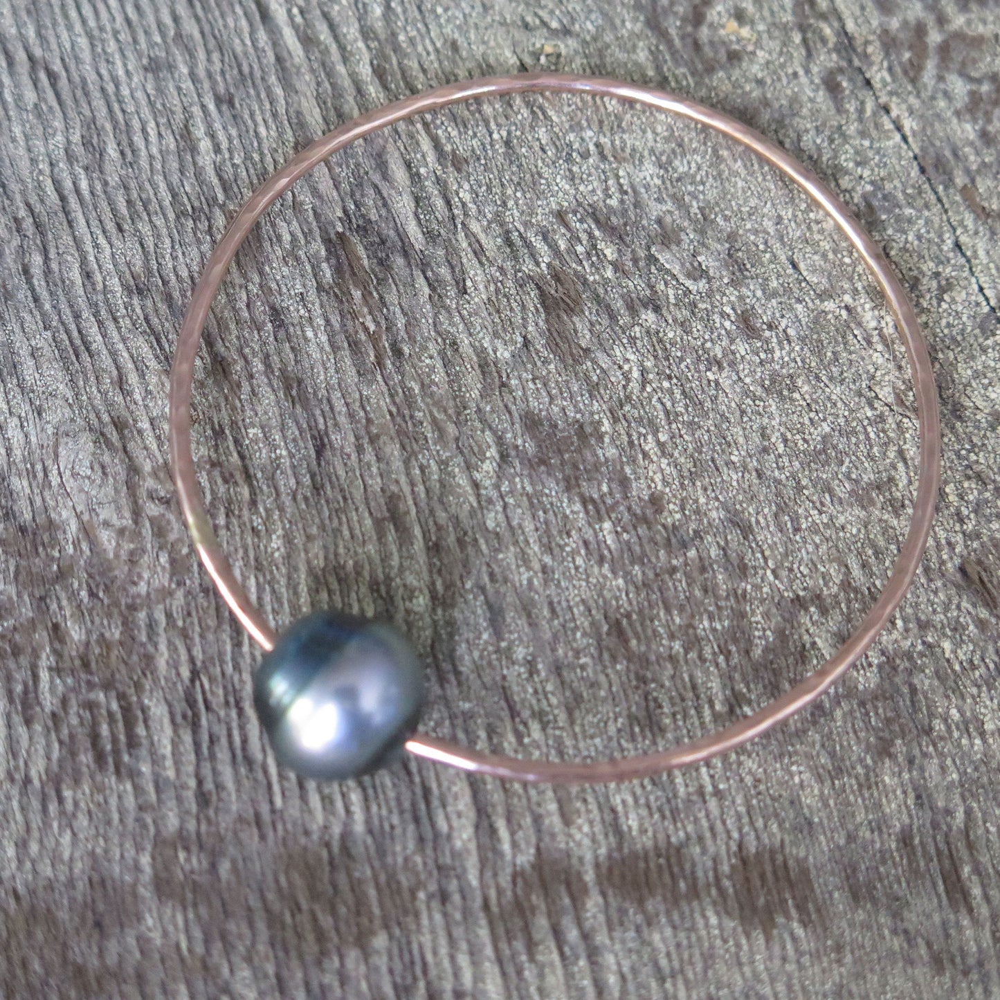 a single black pearl on a rose gold wire shaped into a bangle sits on a wooden background