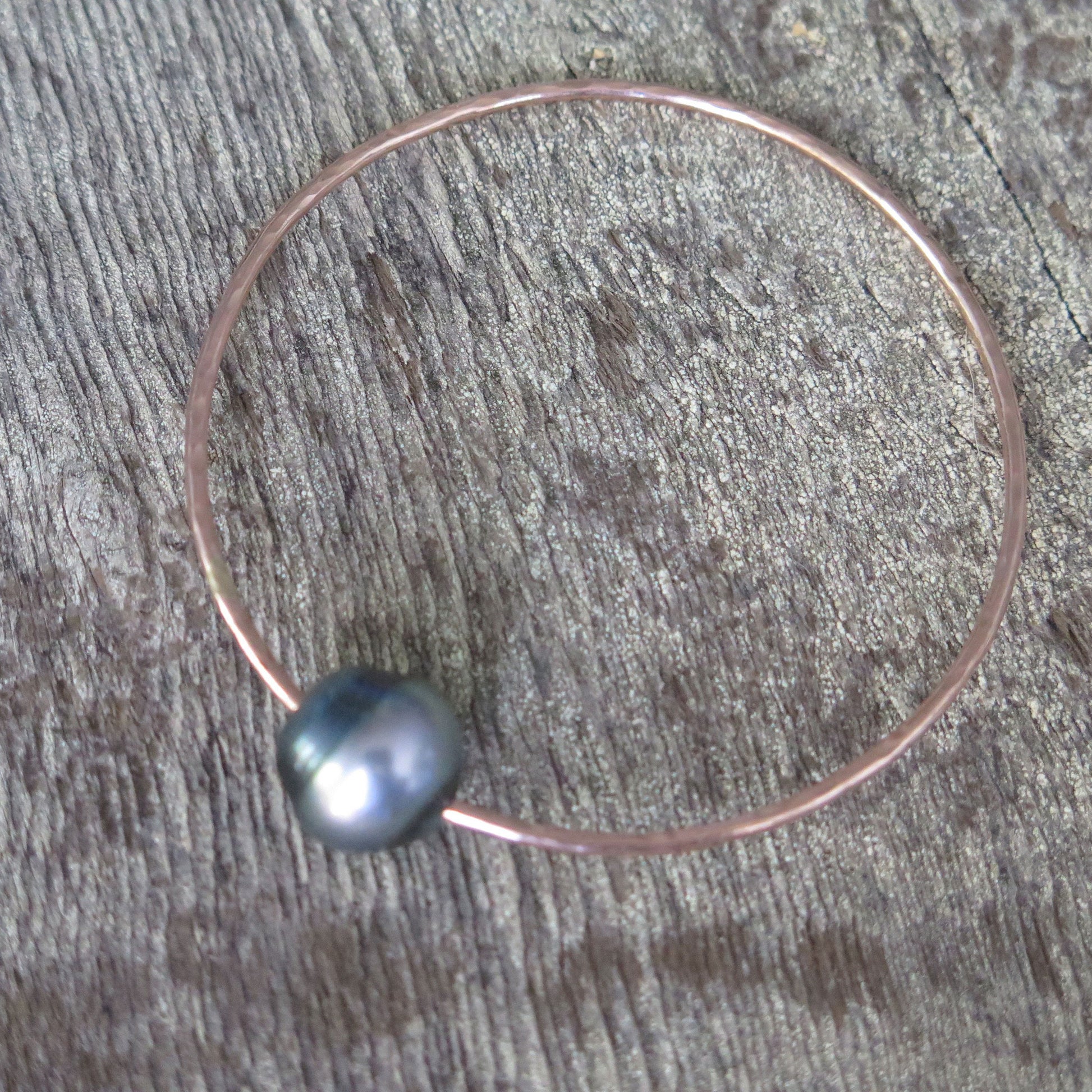 a single black pearl on a rose gold wire shaped into a bangle sits on a wooden background